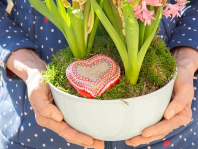 Blumentopf mit einen herzförmigen bemalten Stein, gehalten von zwei Händen.