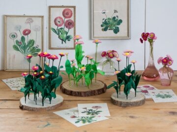 Kunstvolle Papierblumen und echte Margeriten in rosa Glasvasen vor einer Wand mit botanischen Illustrationen.