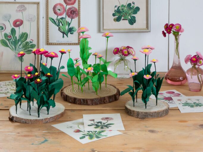 Kunstvolle Papierblumen und echte Margeriten in rosa Glasvasen vor einer Wand mit botanischen Illustrationen. | © Birgid Allig 