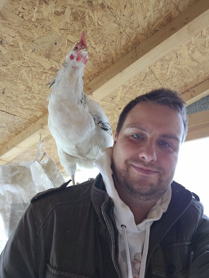 Ein weißes Sussex Huhn sitzt auf der Schulter eines Mannes mit Bart, kurzen Haaren. | © Mattias Nemeth